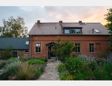 Backsteinhaus mit Satteldach in ländlicher Umgebung, umgeben von einem gepflegten Garten mit wilden Blumen, Sträuchern und einem gepflasterten Weg, der zum Eingangsbereich mit einer überdachten Tür führt. Im Hintergrund sind Bäume sowie ein Anbau mit dunklem Dach erkennbar.