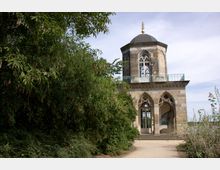 Ein historisches Gebäude mit einer Kuppel und gotischen Fenstern sowie Verzierungen, umgeben von dichter Vegetation. Im Vordergrund verläuft ein sandiger Weg, während das Gebäude unter anderem eine Balustrade und eine Treppe aufweist.