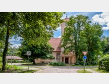 Kirche mit hellem Putz und rotem Ziegeldach, umgeben von großen grünen Bäumen und einer gepflasterten Straße im Vordergrund. Rechts stehen Verkehrsschilder, darunter ein Kreisverkehrszeichen und ein Vorfahrtsschild.