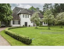 Großes, weißes Landhaus mit dunkelbraunem Schindeldach, umgeben von gepflegtem Rasen, blühenden Bäumen und einer Hecke, die einen Weg entlangführt. Das Gebäude liegt in einer bewaldeten Umgebung mit dichtem Baumbestand im Hintergrund.