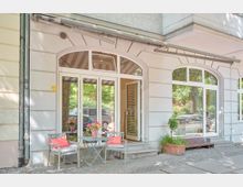 Straßenfassade eines Gebäudes mit bodentiefen, weißen Glastüren und bogenförmigen Fenstern. Im Vordergrund ein kleines Café-Setup mit einem Tisch, zwei Stühlen und Blumen auf einem gepflasterten Gehweg.