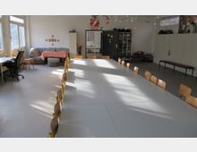 Ein heller Mehrzweckraum mit einer langen Tafel aus zusammengeschobenen Tischen und umstehenden Holzstühlen, seitlich Fenster mit Tageslicht. Im Hintergrund befinden sich eine Sofaecke mit Tisch, Schränke, Regale und Dekoration an der Wand.