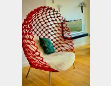 Ein moderner Innenraum mit einem roten Flechtsessel auf schlanken Metallbeinen, einer weißen Polsterung und einem grünen Kissen mit Blattmuster. Im Hintergrund befindet sich ein minimalistisch gestalteter Kamin mit Glasabdeckung und Dekorationen sowie an der Wand kunstvolle Baum-Silhouetten.