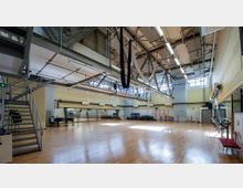 Großer Trainingsraum mit Holzboden, hohen Decken und großen Fenstern, die viel Tageslicht hereinlassen. Der Raum ist mit Spiegelwänden, Turngeräten, einer Metalltreppe sowie hängenden Tüchern und Kletterseilen ausgestattet.