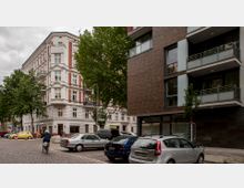 Straßenecke mit einem historischen Gebäude in Weiß und Rot sowie einem modernen Wohnhaus mit glatten Fassaden und Balkonen. Im Vordergrund parken Autos auf einer gepflasterten Straße, während ein Fahrradfahrer vorbeifährt; im Hintergrund sind Bäume sichtbar.