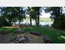 Waldige Uferlandschaft mit einer Feuerstelle aus Steinen und Holzstämmen als Sitzgelegenheiten im Vordergrund, daneben eine Sitzgruppe aus Holztischen und -stühlen. Im Hintergrund ein Steg, der in einen See führt, umgeben von Bäumen.