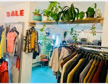 Ein Secondhand-Laden mit Kleidung auf Kleiderstangen und Regalen, umgeben von Zimmerpflanzen auf einem Holzregal und in der Umgebung. Im Hintergrund Fenster, eine Holzstuhlsitzgelegenheit und Dekorationen wie eine Discokugel und Retro-Radios.