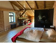 Großer Dachgeschossraum mit freiliegenden Holzbalken und einem Holzboden. Im Vordergrund steht ein Bett mit weißer Bettwäsche und einer roten Decke, im Hintergrund befinden sich schwarze Fliesenwände, ein Fenster mit schwarzem Rahmen und diverse Möbel, darunter ein Regal und eine Leiter.
