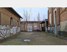Verlassener Industriehof zwischen Backsteingebäuden mit zugemauerten Fenstern und hohen Mauern, die mit Graffiti bedeckt sind. Der Betonboden zeigt Moos und Laub, im Hintergrund stehen kahle Bäume vor einem grauen Himmel.