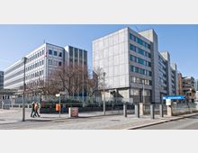 Gebäudeensemble mit moderner, funktionaler Architektur in einer städtischen Umgebung. Im Vordergrund befindet sich die U-Bahn-Station mit dem Schild „U Mohrenstraße“ sowie ein breiter Gehweg mit Passanten.