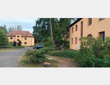 Wohnsiedlung mit zwei hellgelben Gebäuden, die rote Ziegeldächer haben und von Bäumen, Sträuchern und gepflasterten Wegen umgeben sind. Im Vordergrund sind vereinzelt Autos und ein Motorrad zu sehen.