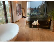 Modernes Schlafzimmer mit freistehender weißer Badewanne und großem Fenster, das den Blick auf Häuser und Bäume draußen bietet. Der Raum hat Holzboden, Dekokerzen auf einem kleinen Tisch und ein Glaswand-Design, das den Schlafbereich abgrenzt.