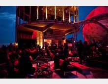 Außenbereich einer alternativen Veranstaltungslocation bei Dämmerung mit mehreren Personen, Sitzmöbeln und Dekorationen. Im Hintergrund sind ein halboffenes Gebäude mit Graffiti und ein beleuchteter Kuppelbau sichtbar.