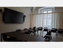 Besprechungsraum mit U-förmig angeordneten dunklen Tischen und Stühlen, ausgestattet mit mehreren Kameras, Mikrofonen und einem Laptop für Videokonferenzen. An der Wand hängt ein großer Bildschirm; gegenüber befindet sich ein großes Fenster mit halbtransparenten Vorhängen.