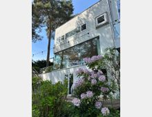 Mehrstöckiges, modernes Wohnhaus mit weißer Fassade, großen Glasfronten und schmalen Fensterbändern, umgeben von hohen Kiefern. Im Vordergrund liegen ein Holzdeck und blühende Sträucher; eine Lichterkette verläuft entlang der Fassade.