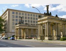 Historische Säulenstruktur mit Verzierungen und Statuen im Vordergrund, dahinter ein großes Verwaltungsgebäude mit klassischer Fassadengestaltung und zahlreichen Fenstern; im Straßenbereich parken mehrere Autos unter blauem Himmel.