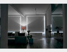 Ein moderner Wohnraum mit großen, abgedunkelten Fenstern, einer grauen Sitzgruppe, einer Stehlampe mit rotem Schirm und mehreren Pflanzen auf kleinen Beistelltischen. Der Raum hat eine minimalistische Einrichtung mit freiliegenden Deckenbalken und hellem Holzboden.