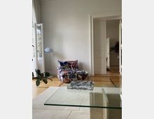 Wohnzimmer mit modernem Design; ein bunter Sessel steht vor einer weißen Wand, daneben eine weiße Stehlampe. Im Vordergrund befindet sich ein Glastisch mit Büchern, und im Hintergrund führt eine geöffnete Tür in einen angrenzenden Raum.