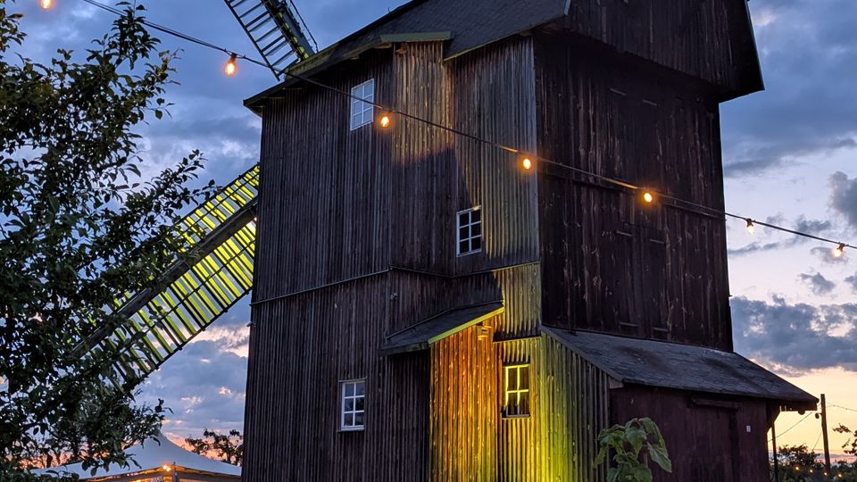 Hölzerne Windmühle mit hohem, dunkel verkleidetem Turm und großen Flügeln, von warmen Lichterketten umspannt, vor einem Abendhimmel mit Wolken. Im Vordergrund stehen Rasenfläche und rustikale Holzelemente, im Hintergrund weitere Gebäude auf dem Gelände.