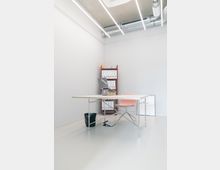 Minimalistisches Büro mit einem weißen Schreibtisch und einer rosa Stuhllehne, einem roten Regal mit Ordnern im Hintergrund, sowie sichtbaren Lüftungsrohren und modernen neonartigen Deckenlampen. Der Raum hat einen hellen Boden, eine weiße Wand und eine schlichte Einrichtung.
