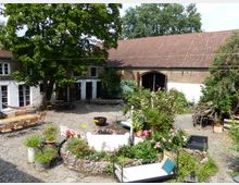 Ein gemütlicher Innenhof mit Kopfsteinpflaster, umgeben von historischen Gebäuden mit Backstein- und weiß verputzten Wänden. Im Hof befinden sich Sitzgelegenheiten, ein Baum, eine kleine bepflanzte Steinmauer, Blumenbeete sowie weitere Dekorationselemente, darunter eine Hängematte und eine offene Feuerstelle.