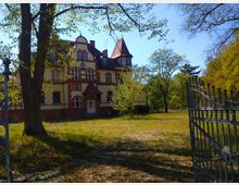 Historisches Gebäude im Stil einer Villa mit gelber Fassade und roten Zierelementen, flankiert von Bäumen in einer grünen Parklandschaft. Im Vordergrund ein geöffneter schmiedeeiserner Torbogen.