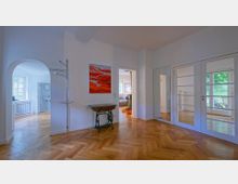 Innenansicht einer hellen Wohnung mit Fischgrätenparkett, weißen Wänden und großen Türen mit Glaseinsätzen. Im Vordergrund ein kleiner Tisch, ein an der Wand befestigter Garderobenständer und ein farbiges Wandbild; im Hintergrund Zimmer mit Fenstern und Blick auf einen grünen Außenbereich.