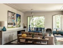 Modernes Wohnzimmer mit beigefarbenem Sofa, dekorativem Couchtisch und großen Wandbildern. Im Hintergrund ein Sitzbereich mit blauen Polstern, großen Fenstern und Blick auf grüne Bäume.