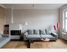 Helles Wohnzimmer mit grauem Sofa, weißem Kamin, Holzfußboden und einer Backsteinwand auf der linken Seite. Vor dem Sofa steht ein kleiner rosafarbener Beistelltisch, eine Stehlampe und ein Hund liegt auf dem Boden.