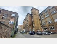 Backstein-Industriegebäude aus dem späten 19. oder frühen 20. Jahrhundert, umgeben von einer Kopfsteinpflasterstraße. Links eine Wand mit Graffiti, rechts geparkte Autos vor weiteren historischen Gebäuden mit großen Fenstern und Rundbogenarchitektur.