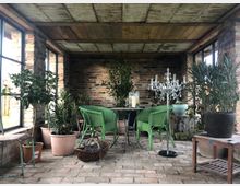 Ein kleiner Wintergarten mit Backsteinwänden, großen Fensterfronten und Holzbalkendecke. Der Raum ist mit grünen Rattan-Stühlen, einem schwarzen Metalltisch, zahlreichen Pflanzen in Töpfen und einem dekorativen Kristall-Kronleuchter gestaltet.