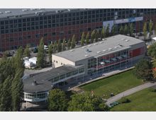 Luftaufnahme eines modernen Gebäudekomplexes mit Flachdach und großen Glasfronten. Im Vordergrund ein Rundbau mit verglastem Obergeschoss, umgeben von Bäumen und einer Grünfläche, im Hintergrund ein mehrstöckiges Bürogebäude mit roten und grauen Fassadenelementen.