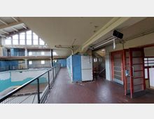 Innenansicht eines alten, verlassenen Schwimmbads mit einer leeren Halle und abgenutzten Umkleidekabinen. Die Umgebung zeigt Fliesenwände, Metallgeländer und Fenster mit viel Tageslicht sowie beschädigte Oberflächen.