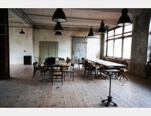 Industrie-inspirierter Besprechungsraum mit Holzboden, einem langen Tisch aus Holz mit verschiedenen Stühlen und großen Fenstern mit Jalousien. An der Decke hängen mehrere schwarze Metalllampen, und die Wände sind teils verputzt, teils aus Sichtmauerwerk.