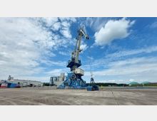 Großer blauer Hafenkran auf einem weitläufigen Betonplatz, daneben Industriegebäude und Silos im Hintergrund. Der Himmel ist bewölkt mit einzelnen klaren blauen Abschnitten.
