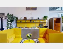 Moderner Gemeinschaftsraum mit gelber Sitzecke, einem kleinen runden Tisch mit einem Blumenstrauß sowie einer Rückwand mit gelben Regalen, dekorativen Gegenständen und einem Buffet mit Speisen und Getränken. Im Hintergrund sind eine große Zimmerpflanze und ein separater Holzarbeitsbereich sichtbar.