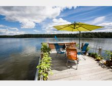 Holzterrasse an einem See mit Blick auf das Wasser und einen bewaldeten Uferbereich im Hintergrund. Auf der Terrasse stehen bunte Gartenstühle um einen runden Tisch, daneben ein großer gelber Sonnenschirm; Pflanzen und Töpfe säumen den Rand der Plattform.