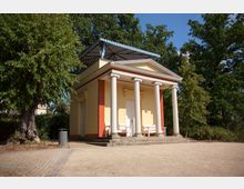 Klassizistisches Gebäude mit gelber Fassade, weißen Säulen und einem Dreiecksgiebel, umgeben von Bäumen. Im Vordergrund befinden sich ein sandiger Platz, Kopfsteinpflaster und eine Sitzbank.
