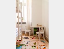 Ein helles Kinderzimmer mit einem Hochbett, einem kleinen weißen Tisch und zwei passenden Stühlen mit sternförmigen Ausschnitten in den Rückenlehnen. Der Boden ist mit einem bunten Teppich mit Tier- und Fahrzeugmotiven bedeckt, während das Tageslicht durch große Fenster mit weißen Vorhängen einfällt.
