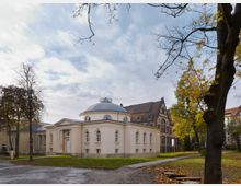 Freistehendes Gebäude mit Rundkuppel und klassizistischen Elementen, umgeben von herbstlichen Bäumen; im Hintergrund ein größeres Backsteingebäude mit schrägem Giebel.
