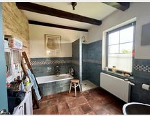 Rustikales Badezimmer mit freiliegenden Holzbalken, wandhängender Badewanne und ebenerdiger Dusche mit blaugrauen Fliesen. Eine Fensterbank mit Rollos, ein Waschbecken aus Stein und antik wirkende Holzmöbel vervollständigen das Ambiente.