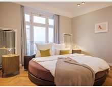 Modernes Schlafzimmer mit einem runden Bett, weißen Kissen und einer beigen Tagesdecke. Bodentiefe Fenster mit grauen Vorhängen, zwei Nachttische mit goldenen Lampen und ein minimalistisches Wandbild runden den Raum ab.