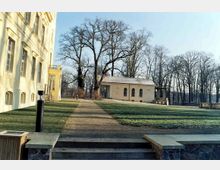 Historischer Gebäudekomplex mit gelb gestrichener Fassade auf der linken Seite, angrenzend an einen gepflegten Park mit Rasenflächen und einem kleinen Pavillon im Hintergrund, umgeben von kahlen Bäumen im Winter.