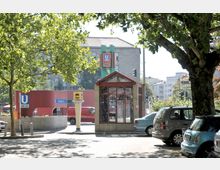 Eingangsbereich einer U-Bahn-Station mit roter Überdachung und einem Schild, das auf die Linien U3 und U7 hinweist, umgeben von Bäumen, geparkten Autos und einem Taxistand. Im Hintergrund sind ein mehrstöckiges Gebäude und weitere Straßenelemente zu sehen.