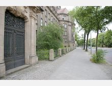 Historisches Gebäude mit verzierten Toren und steinernen Fassaden neben einem Gehweg, der von Bäumen gesäumt ist. Im Hintergrund verläuft eine Straße mit weiteren Bäumen und einigen parkenden Autos.