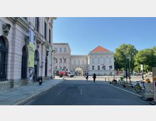 Straßenszene mit historischen Gebäuden, darunter ein rosafarbenes Gebäude mit großen Fenstern und Bannern sowie ein weißes Gebäude mit rotem Dach. In der Mitte befindet sich ein Torbogen, durch den Fußgänger und Radfahrer sichtbar sind. Im Vordergrund sind Fahrräder, Roller und Absperrungen erkennbar, während Bäume den Hintergrund ergänzen.