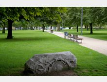 Rasenfläche in einem Park mit Bäumen, entlang eines hellen Kieswegs, der an Sitzbänken und Papierkörben vorbeiführt. Im Vordergrund ein großer steinerner Dekorblock mit eingravierten Mustern und Pflanzenmotiven.