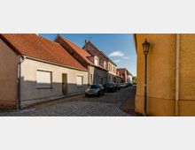 Schmale Kopfsteinpflasterstraße in einem Wohnviertel mit niedrigem Reihenhausbestand und roten Ziegeldächern. Entlang der Straße stehen mehrere geparkte Autos; an der rechten Hauswand befindet sich eine Straßenlaterne.