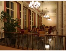 Ein eleganter, heller Innenraum mit großen Fenstern und langen Vorhängen, bestückt mit Tischen und Stühlen. Der Raum wird von mehreren Kronleuchtern beleuchtet, hat eine verzierte Decke und wird von runden, verkleideten Säulen strukturiert.