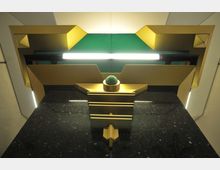 Detailaufnahme eines kunstvollen, symmetrischen metallischen Designs in Gold- und Grüntönen mit geometrischen Formen. Ein leuchtendes Element verläuft horizontal durch die Mitte, darunter befindet sich eine kugelförmige, grüne Verzierung auf einer goldenen Basis.